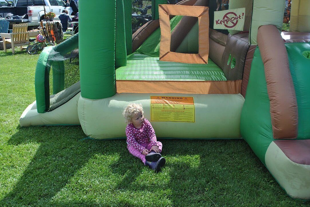 A little girl is sitting on the grass next to a bouncy house.
