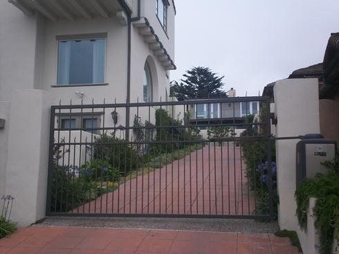 Gated driveway leading to a house with white walls, a red brick path, and greenery along the sides