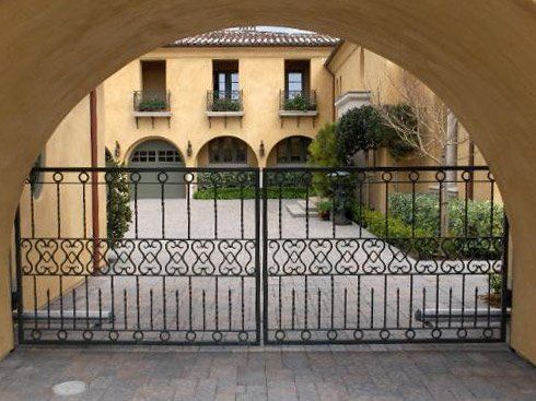 Arched view through a wrought-iron gate toward a cream-colored villa with windows and a garden entrance