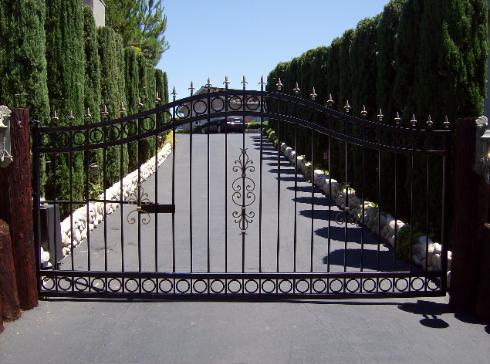 Black wrought-iron driveway gate blocking a paved path lined with tall green trees