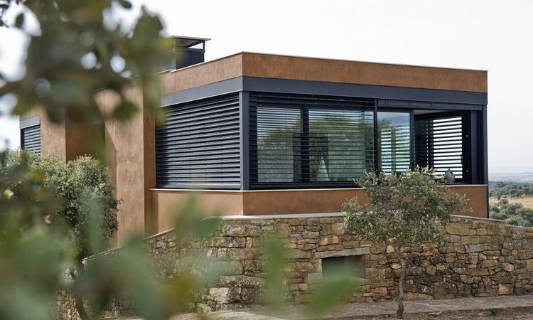 Casa moderna con dettagli in legno, tende orizzontali scure e paesaggio in pietra dietro una vegetazione sfocata.
