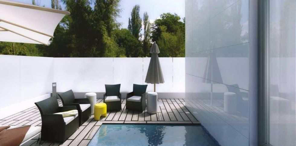 Moderno patio esterno con sedute grigie, cuscini gialli, una piscina e piante alte dietro un muro bianco.
