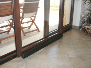 Porta scorrevole in vetro con soglia scura che si apre su un patio con sedie in legno e pavimento piastrellato.