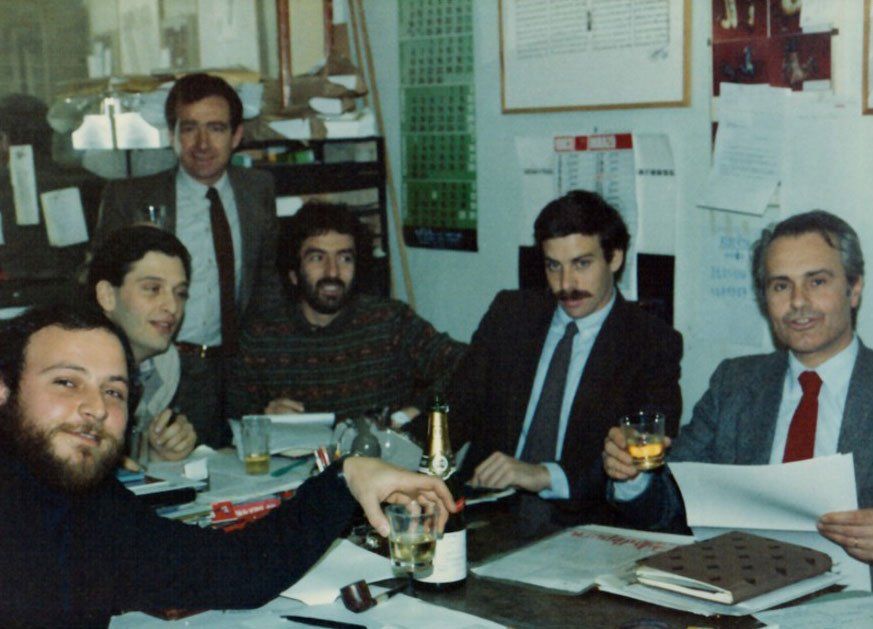Un gruppo di uomini seduti attorno a un tavolo con un calendario sul muro dietro di loro