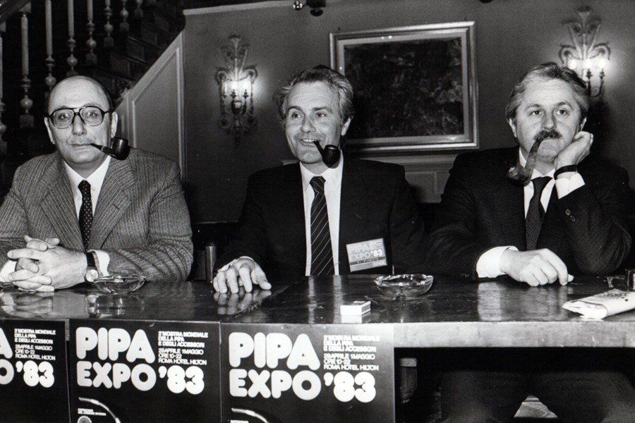 Tre uomini seduti a un tavolo con un cartello che dice pipa expo 83