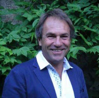 Un uomo con una giacca blu e una camicia bianca sorride davanti a un cespuglio.