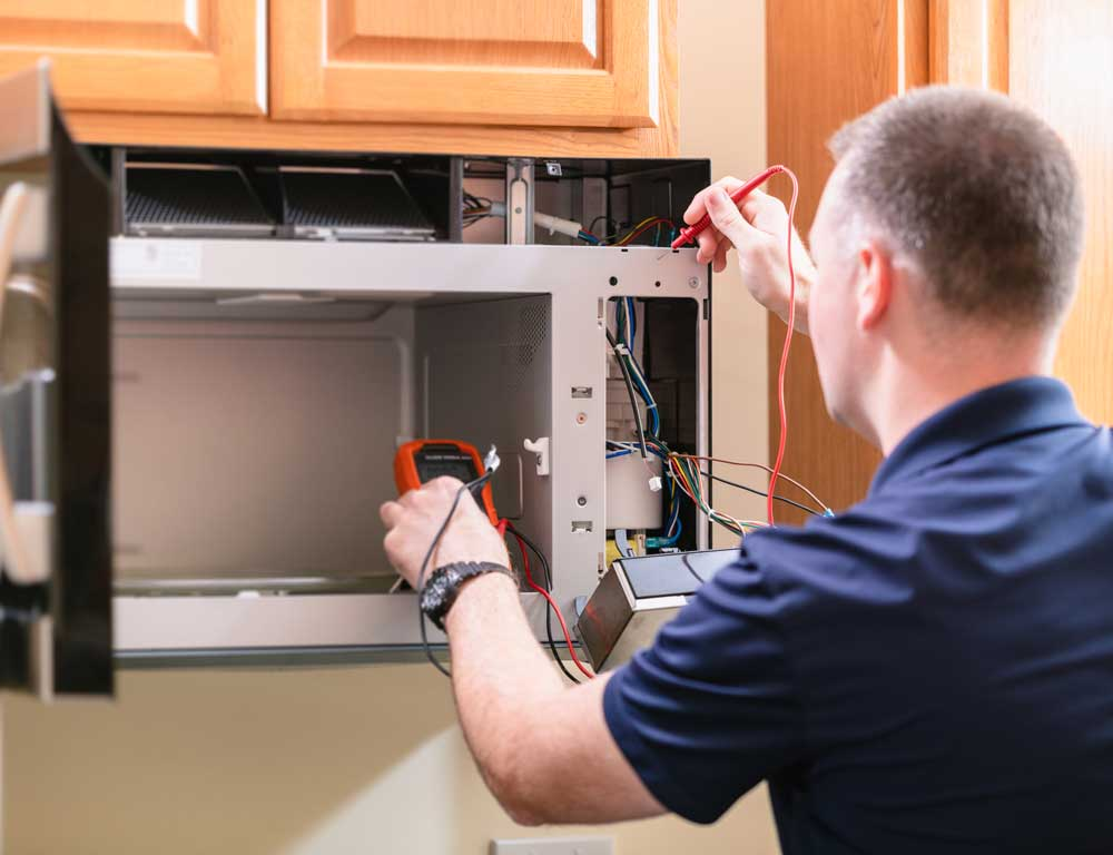 Technician using a multimeter to repair an open microwave under kitchen cabinets