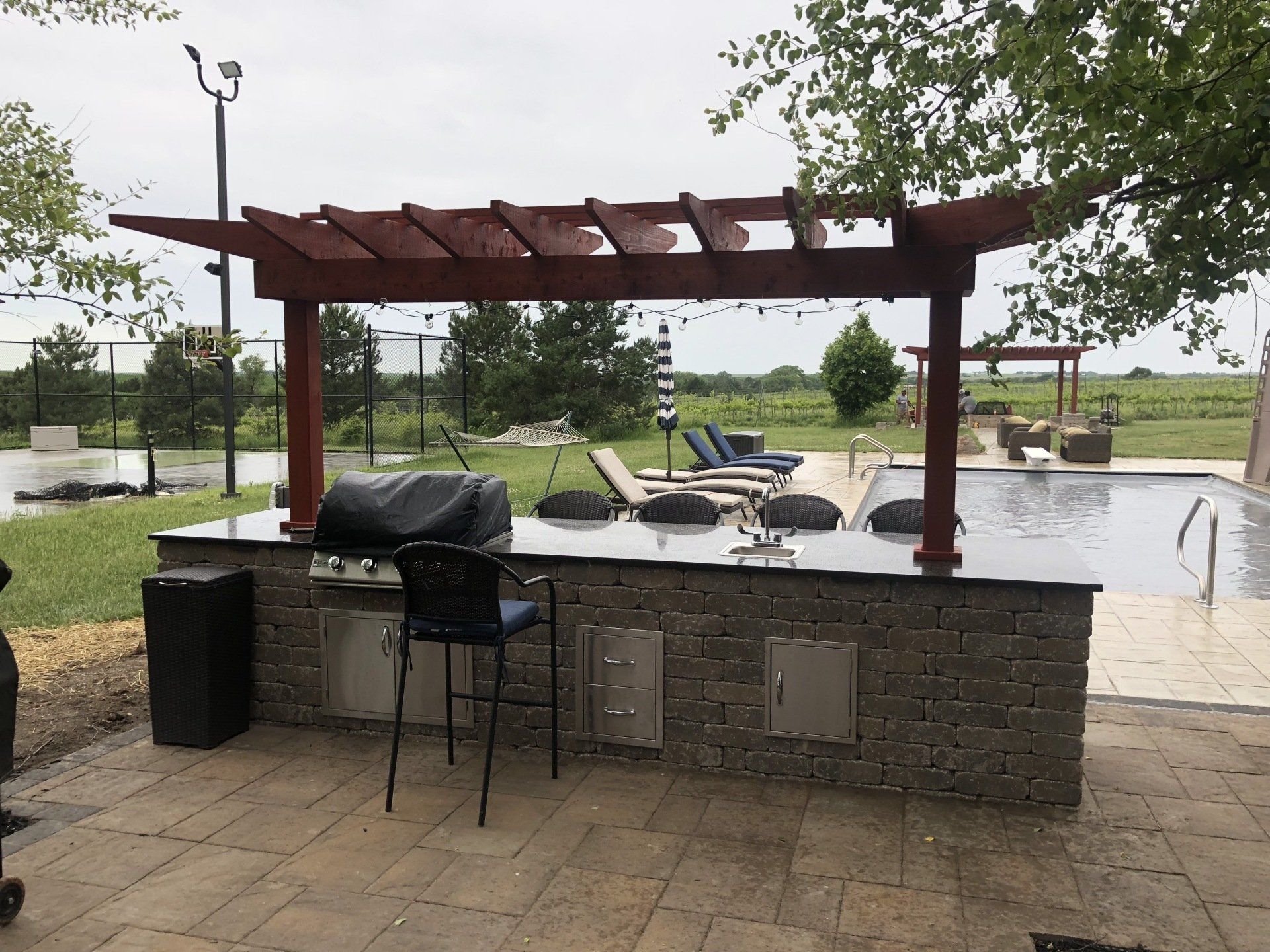 A large outdoor kitchen with a pergola over it and a swimming pool in the background.