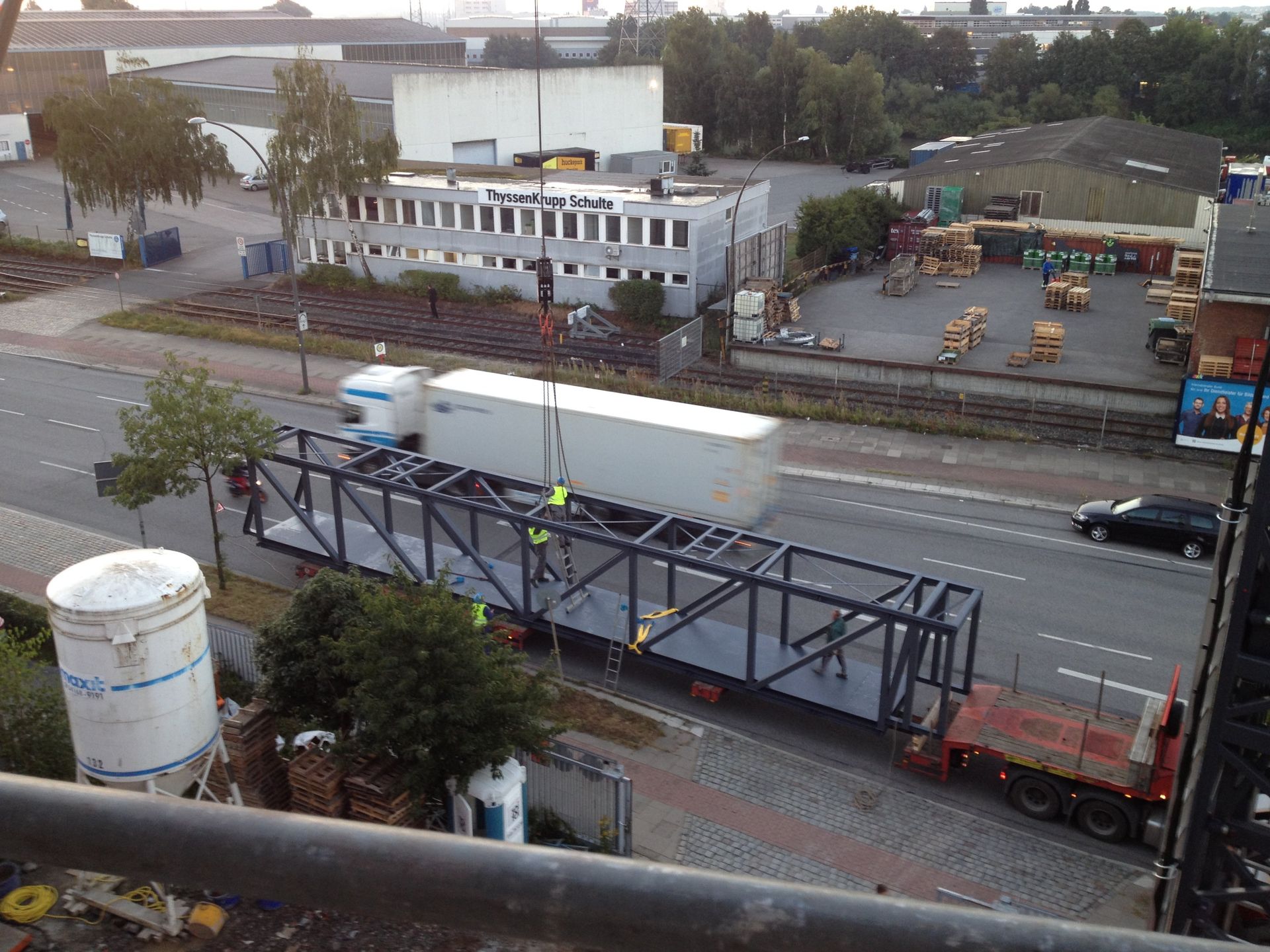 Straßenbaustelle neben einer Straße, ein Kran hebt einen weißen Container in der Nähe eines umzäunten Arbeitsbereichs an.