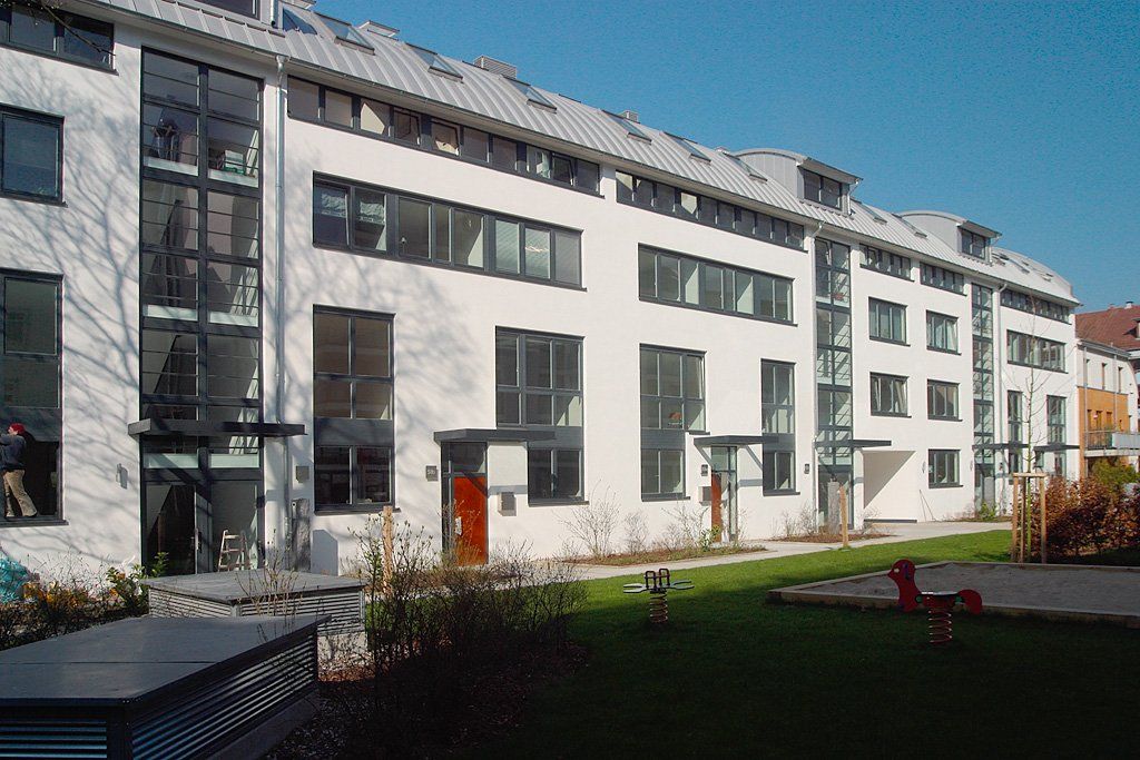 Modernes weißes Wohnhaus mit Balkonen, roten Türen und einem begrünten Innenhof unter blauem Himmel