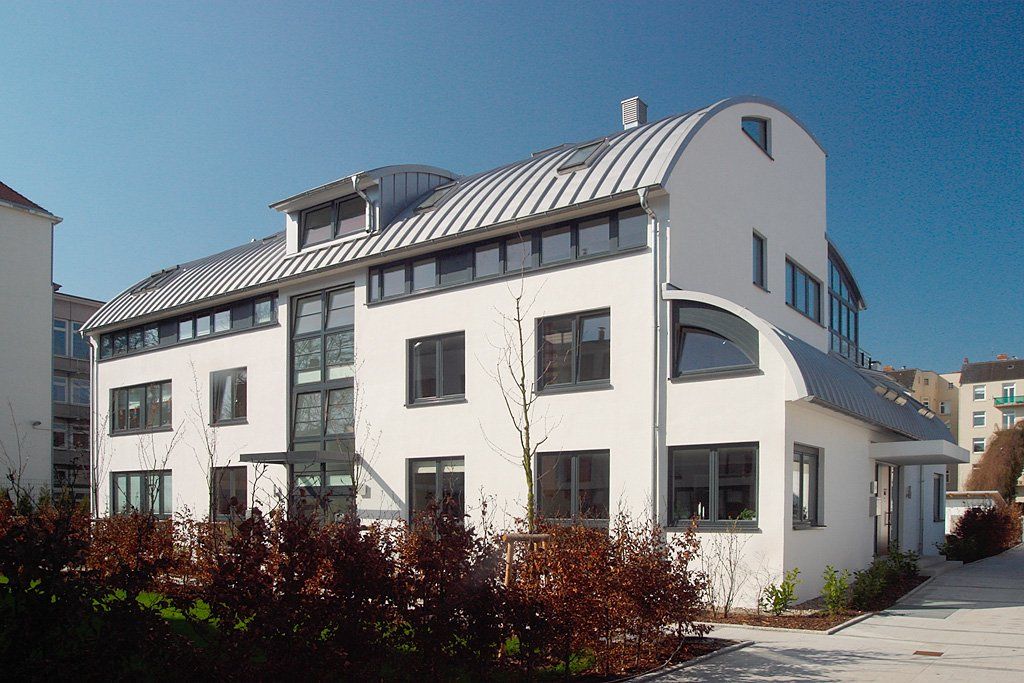 Weißes, modernes Mehrfamilienhaus mit Balkonen und Ziegeldach an einer sonnigen Straße