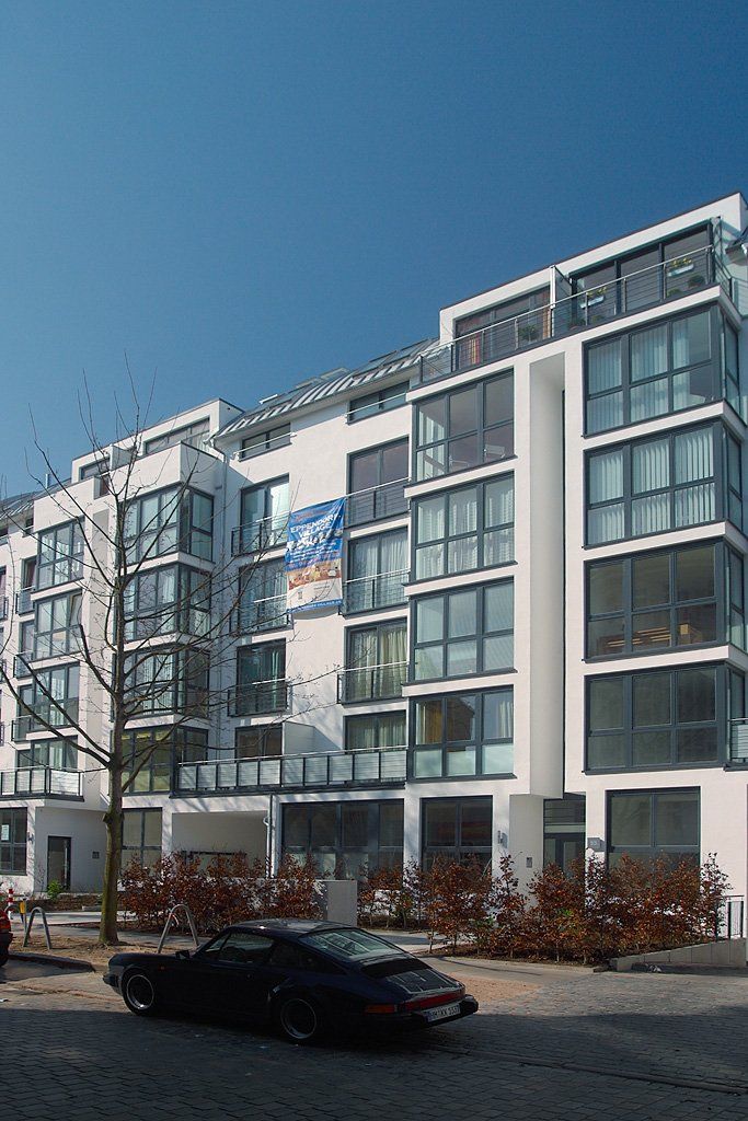 Modernes Mehrfamilienhaus mit Balkonen, großen Fenstern und einem geparkten Auto in einer sonnigen Straße