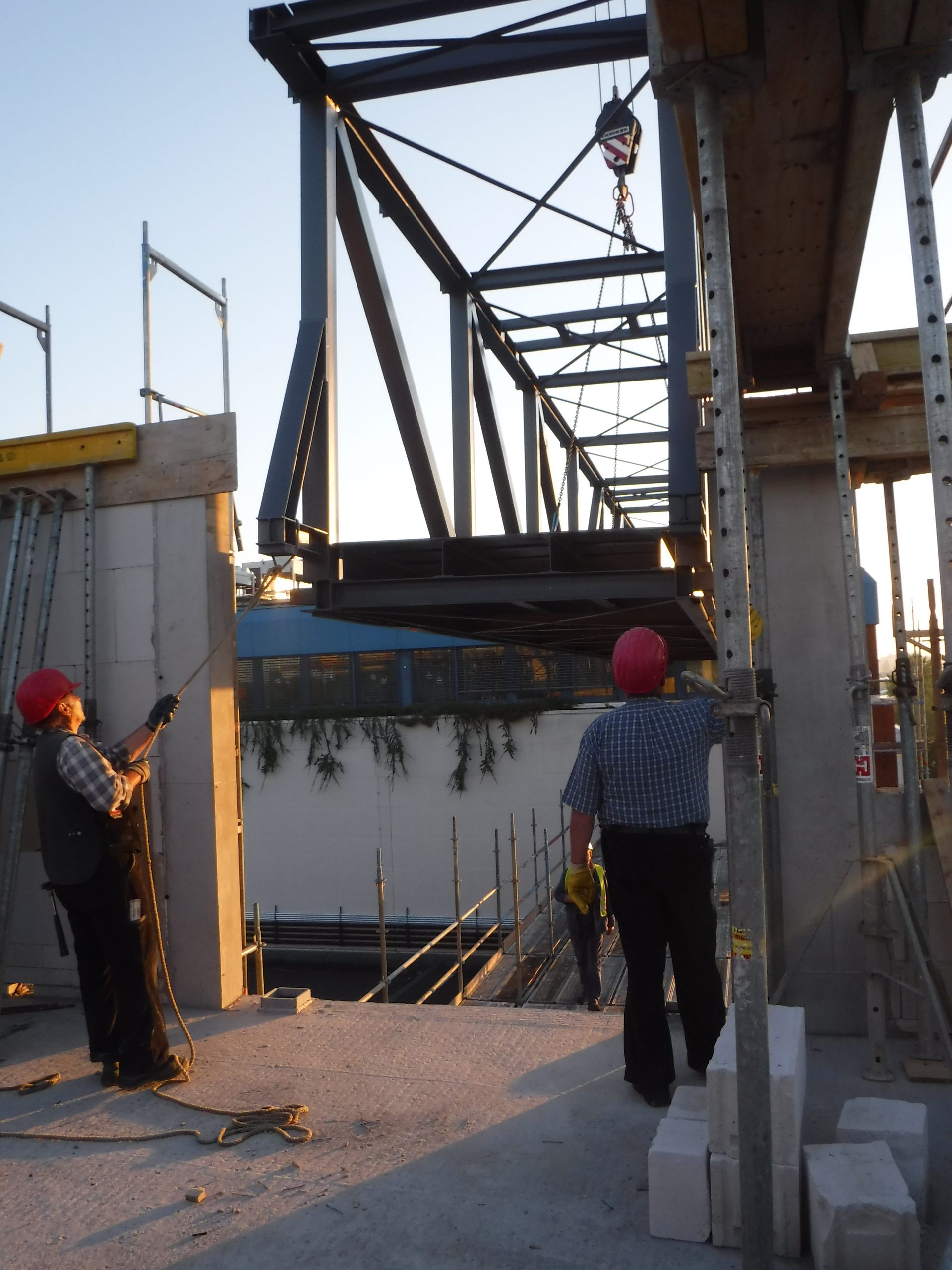 Zwei Arbeiter mit Schutzhelmen stehen neben einem Teilstück einer hängenden Stahlbrücke auf einer Baustelle.
