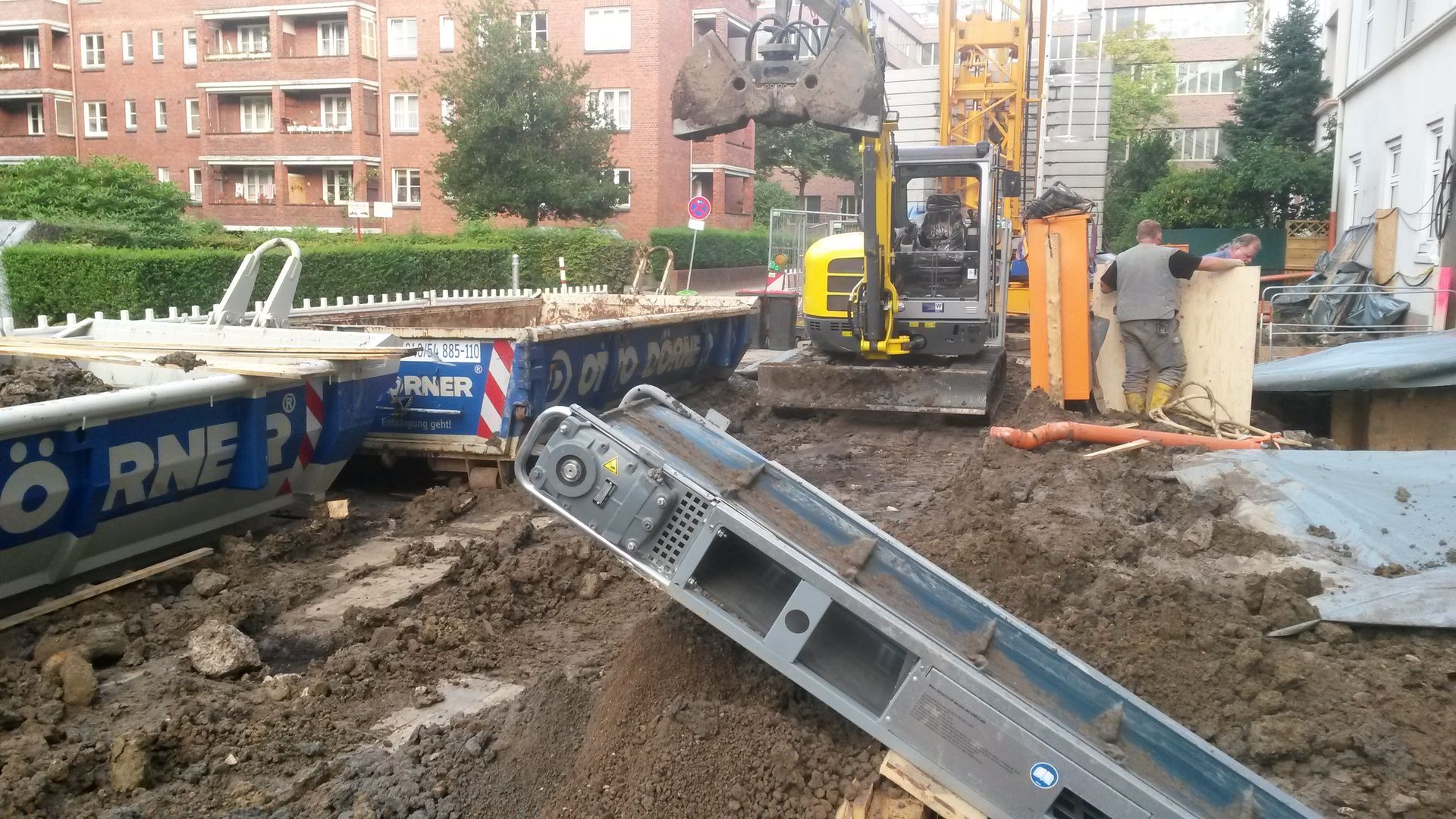 Baustelle mit Bagger und geneigter Metallbarriere über schlammigem Boden in der Nähe von Wohnhäusern