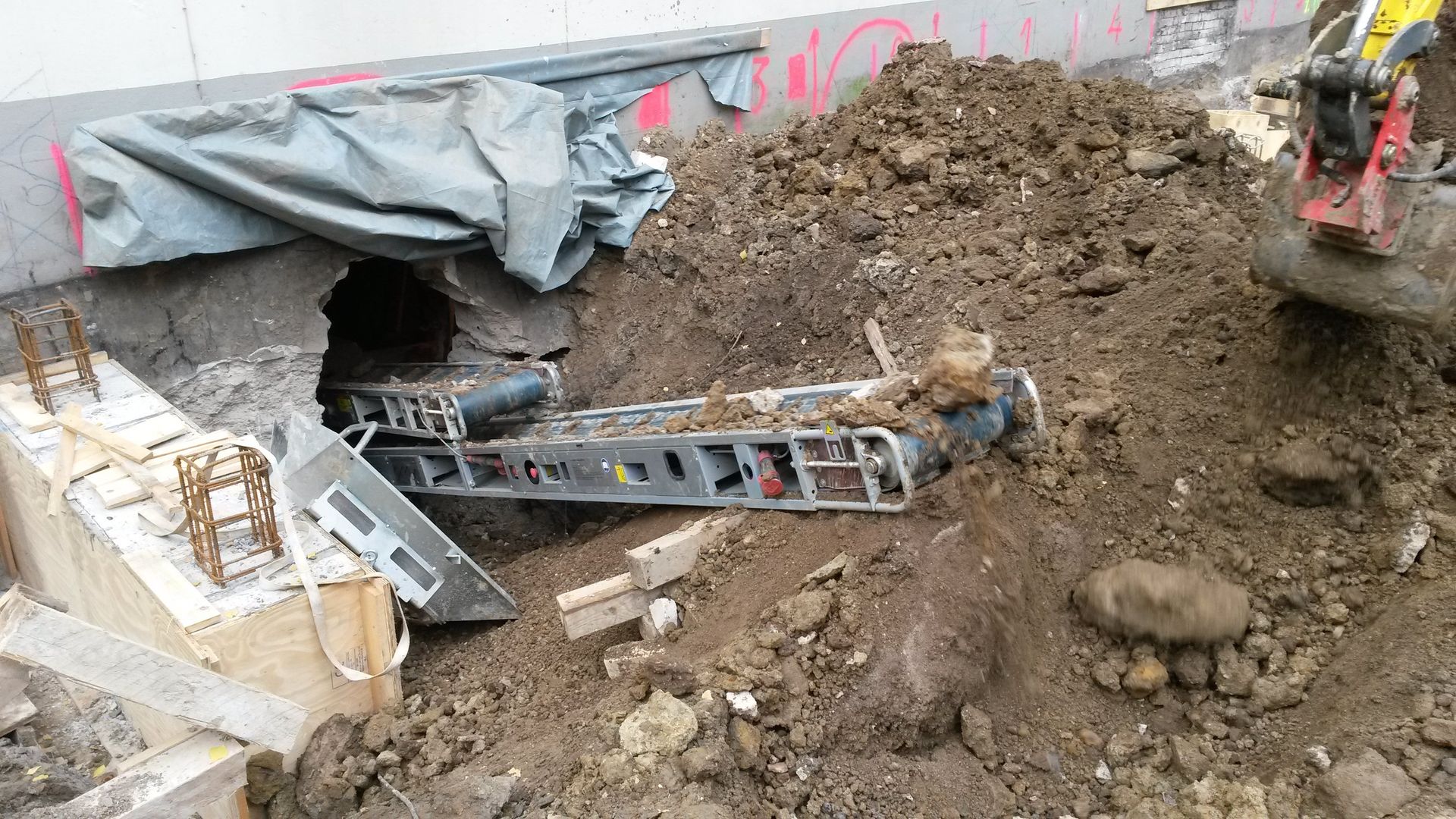 Ein Bagger gräbt neben einem mit einer Plane abgedeckten Loch, um das herum Schutt und Erde aufgeschüttet sind.