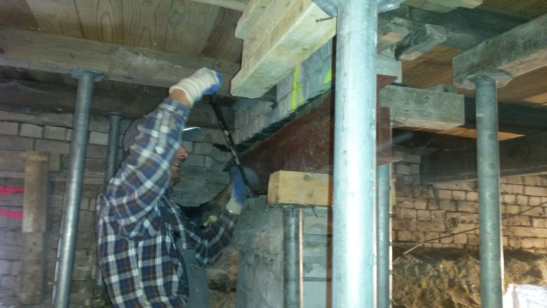Ein Arbeiter in einem Hemd benutzt eine Bohrmaschine unter einem Holzboden in einem Kriechkeller mit Metallstützpfosten.