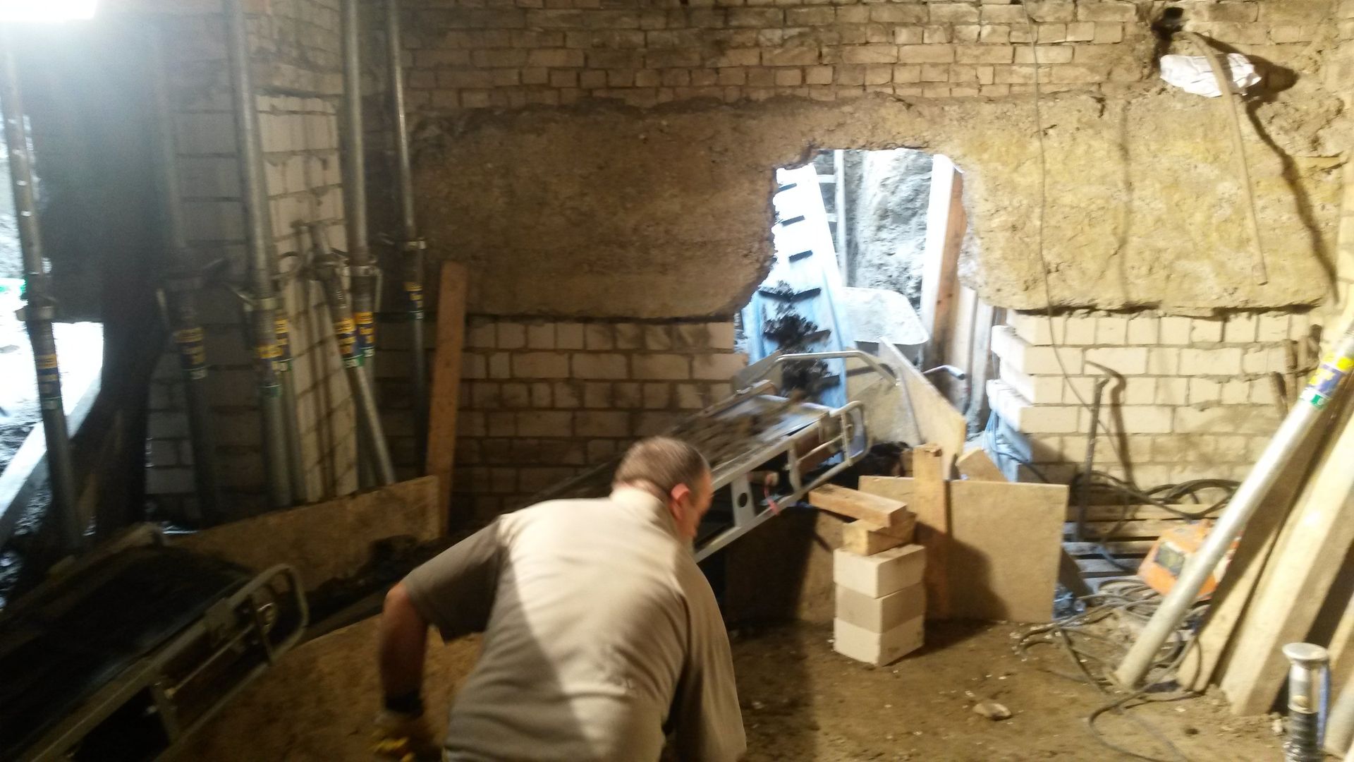 Zwei Arbeiter in einer gemauerten Kellerwerkstatt in der Nähe einer großen Öffnung, umgeben von Werkzeugen und Kisten.