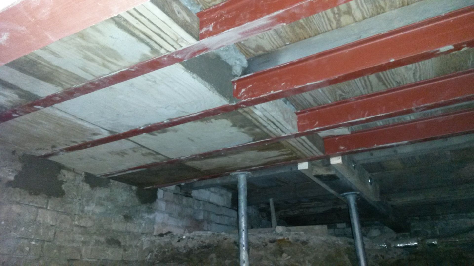 Rissige Betondecke mit roten Balken und metallenen Stützpfeilern in einem dunklen Keller