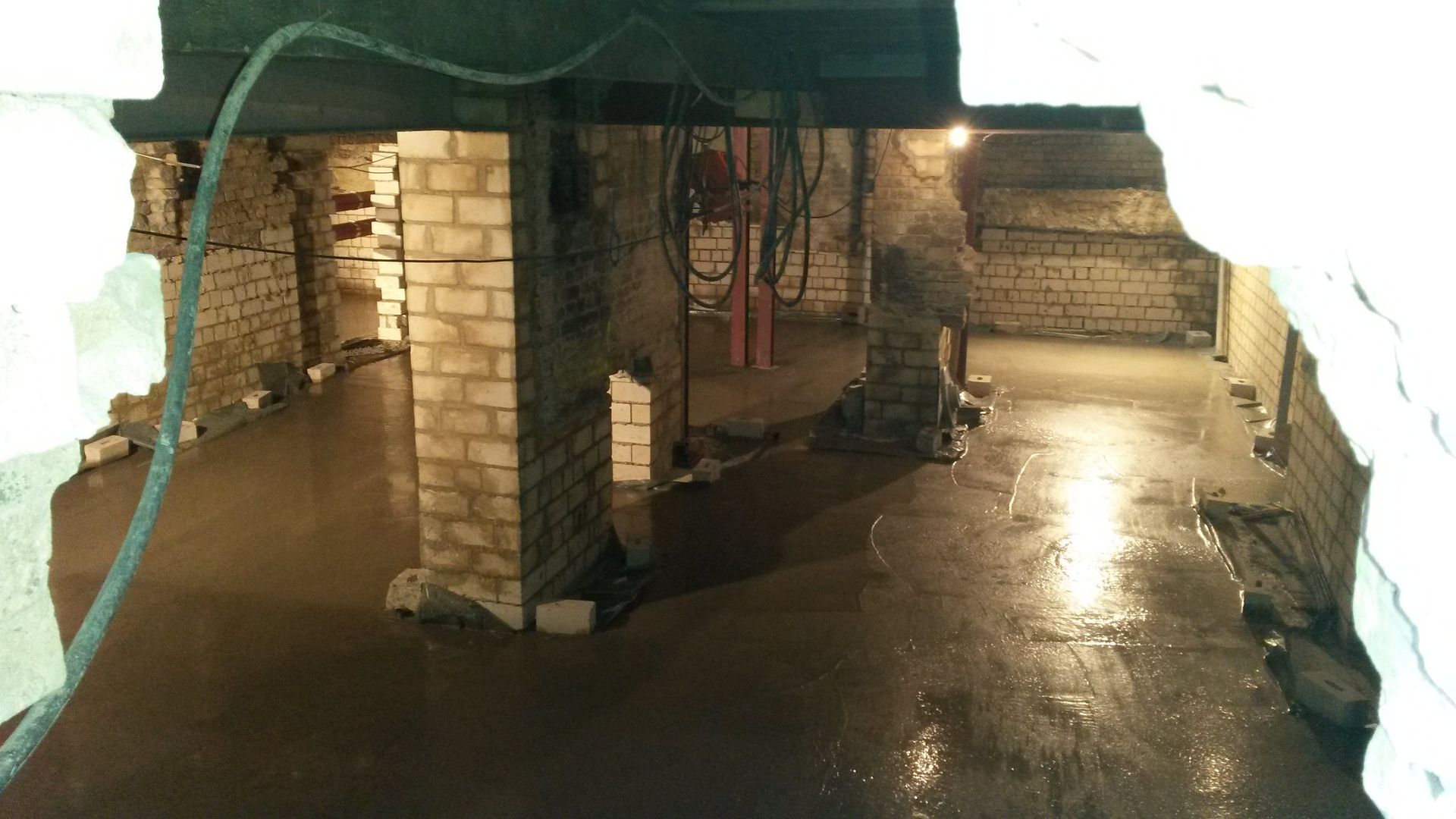 Überfluteter Ziegelkeller mit nassem Betonboden und Stützpfeilern unter einer niedrigen Decke