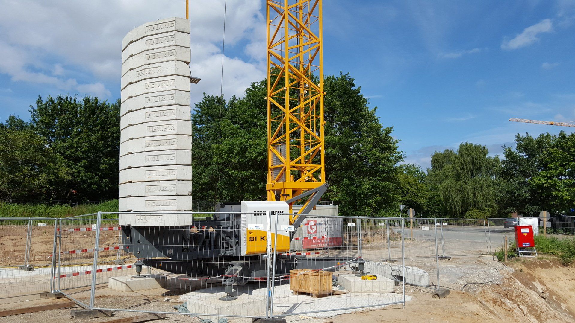 Baustelle mit hohem weißen Silo und gelbem Kran vor Bäumen und blauem Himmel