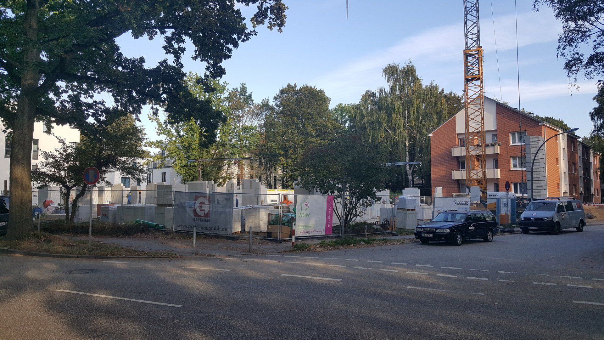 Straßenansicht einer Baustelle mit Bäumen, einem Kran, geparkten Autos und Wohnhäusern im Hintergrund