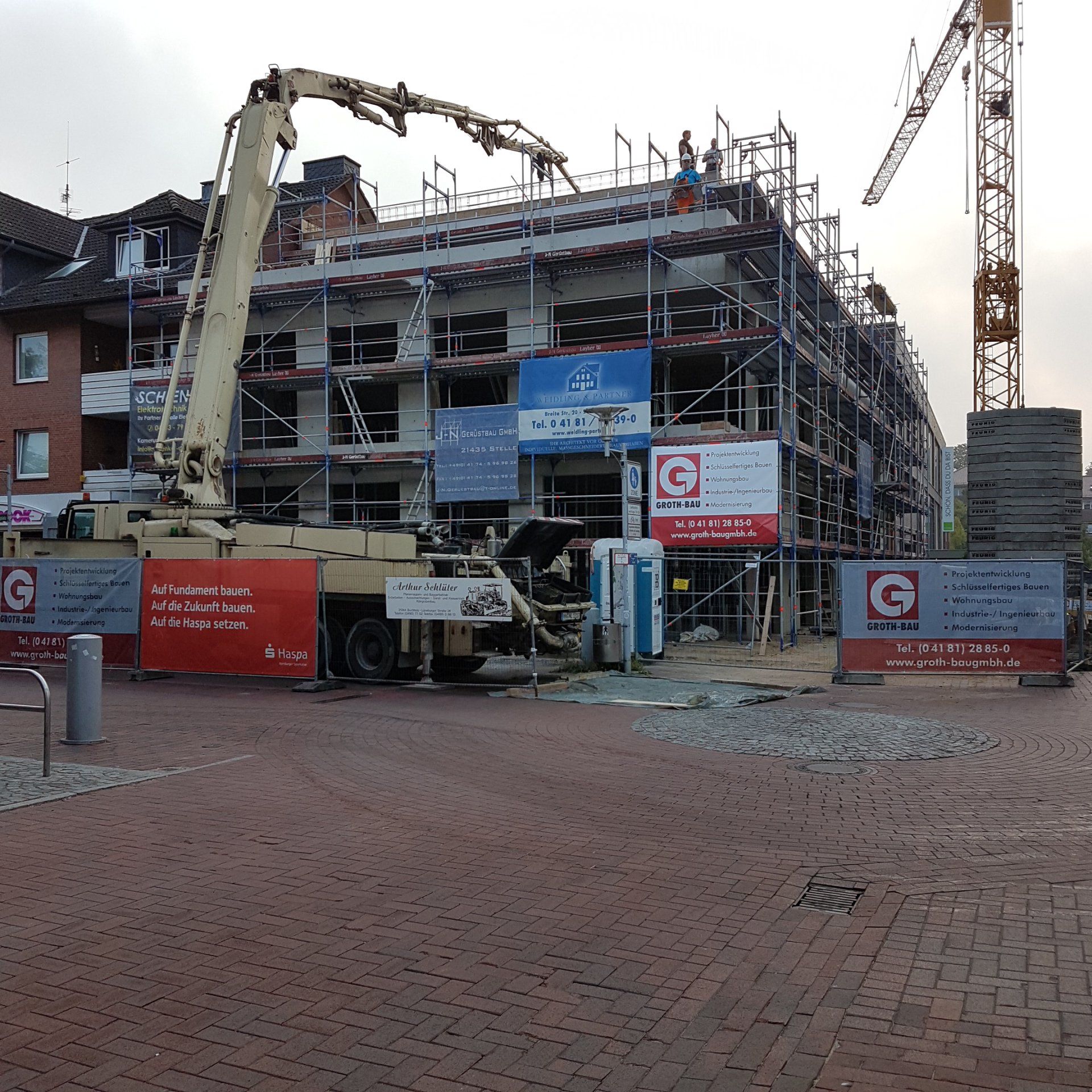 Baustelle mit Kränen, Arbeitern und einem teilweise errichteten mehrstöckigen Gebäude hinter roten Absperrungen.