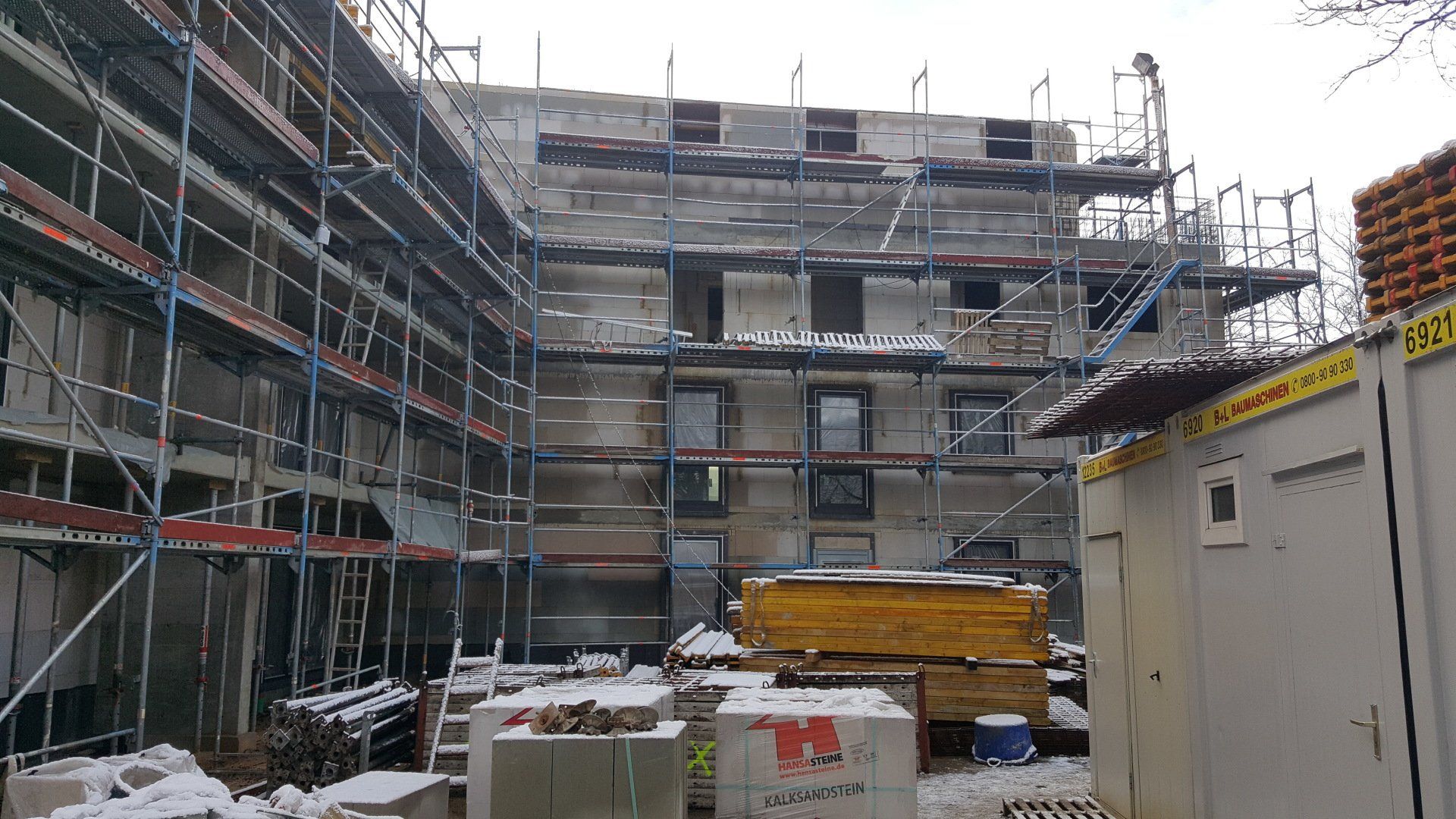 Verschneite Baustelle mit Gerüsten um ein Gebäude und gestapelten Materialien im Vordergrund