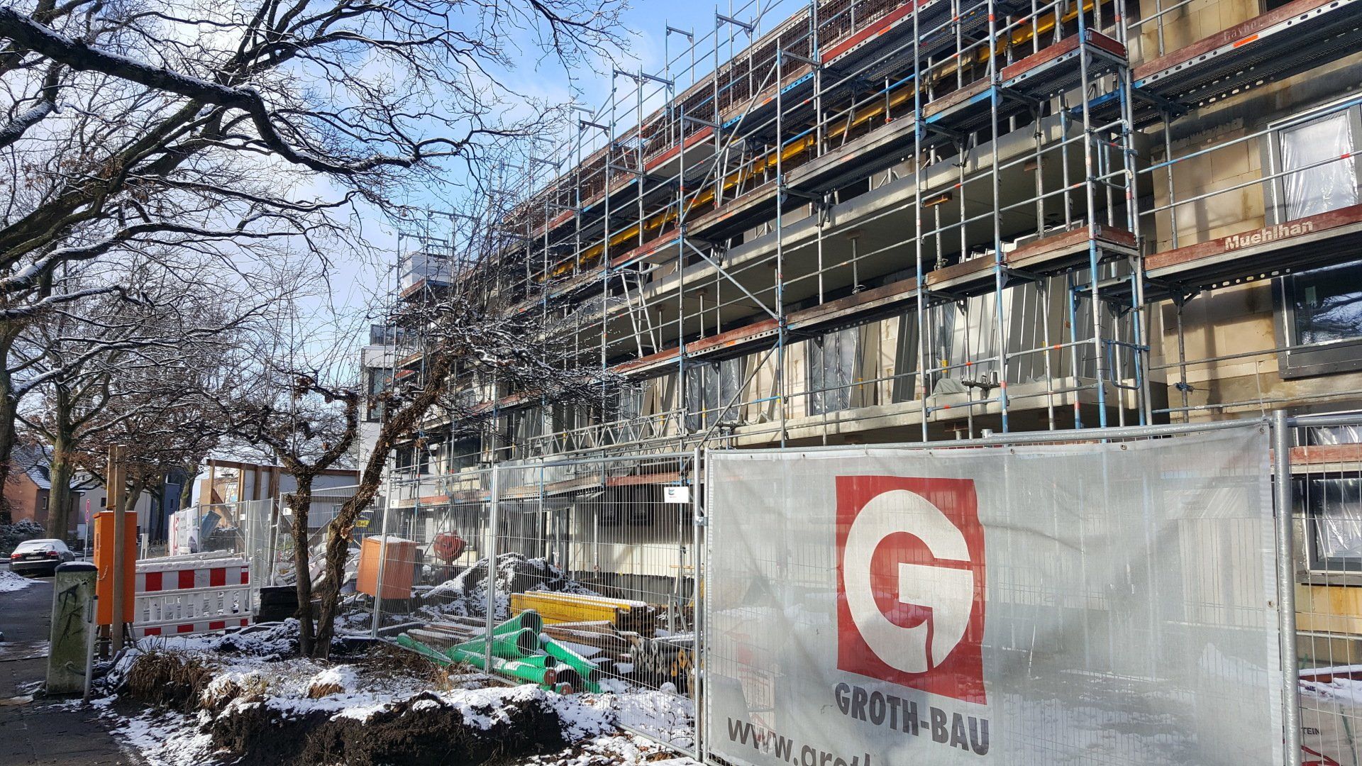 Baustelle mit Gerüst neben einer von Bäumen gesäumten Straße und einem weißen Zaunschild davor