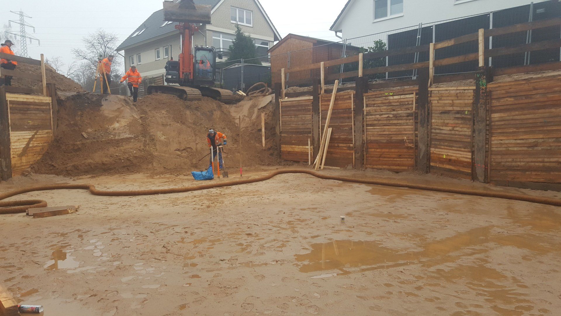 Baustelle mit ausgehobener Erde, hölzernen Stützmauern und Häusern im Hintergrund unter einem bewölkten Himmel
