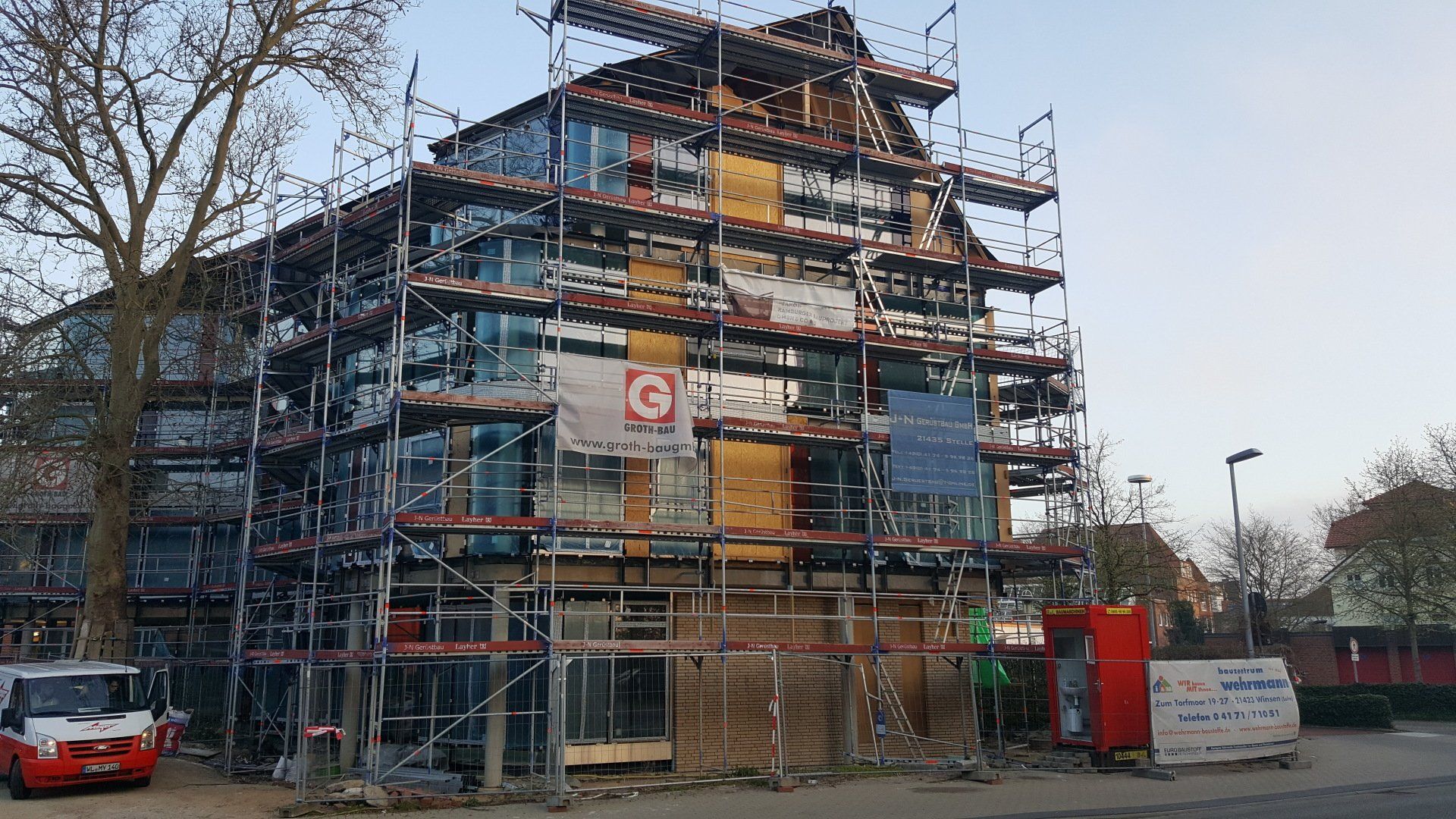 Ein mehrstöckiges, im Bau befindliches Gebäude ist von einem Gerüst umgeben; in der Nähe ist ein rotes Auto geparkt.
