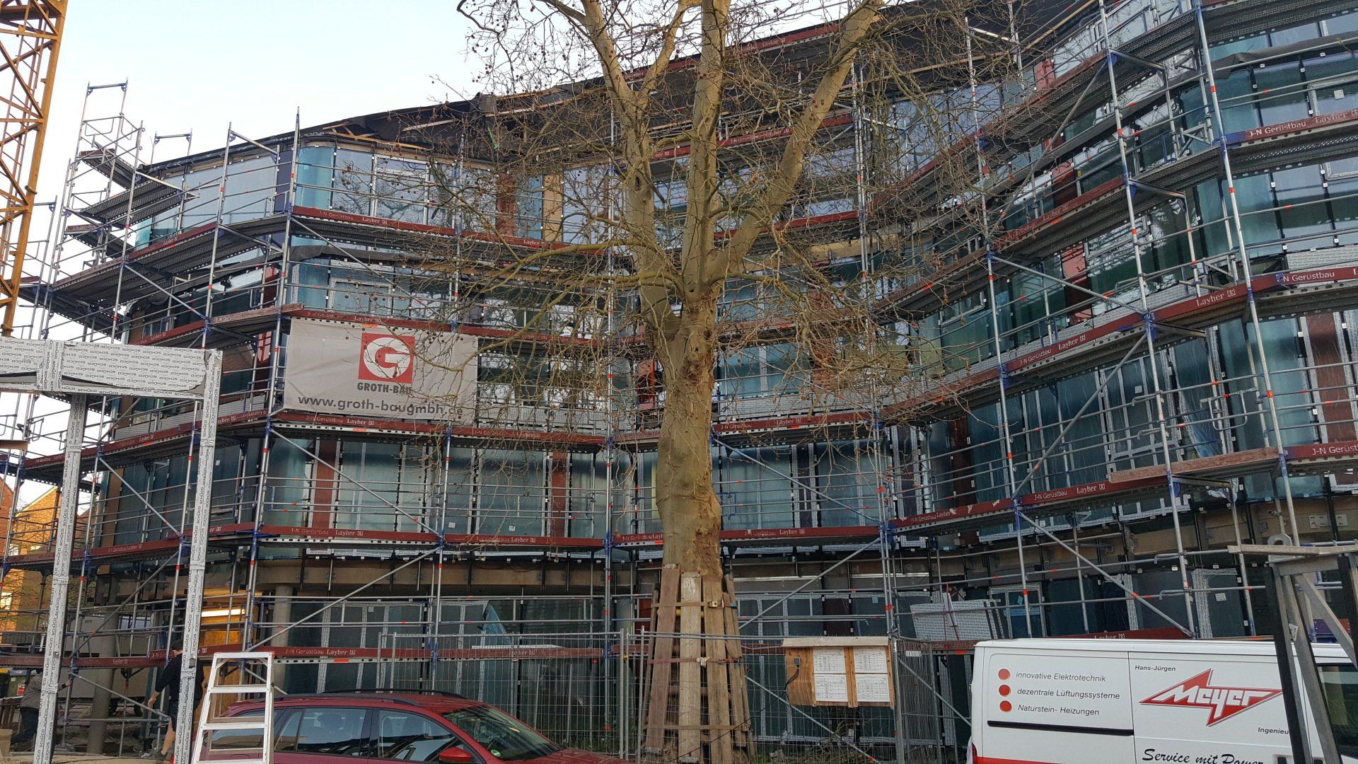 Ein Baugerüst umgibt ein mehrstöckiges, im Bau befindliches Gebäude; davor steht ein kahler Baum.