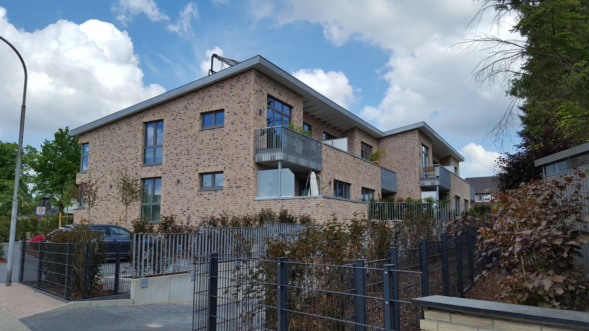 Backstein-Mehrfamilienhaus mit Balkonen hinter einem Metallzaun unter einem teilweise bewölkten Himmel