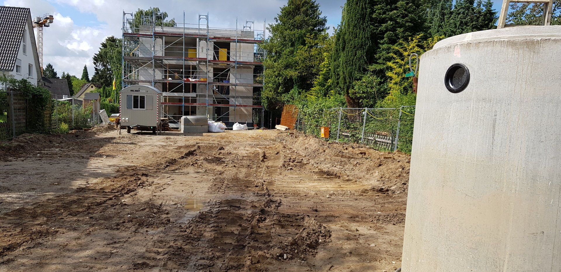Baustelle mit schlammigem Boden, einem teilweise errichteten Haus und einem weißen Betonpfosten im Vordergrund