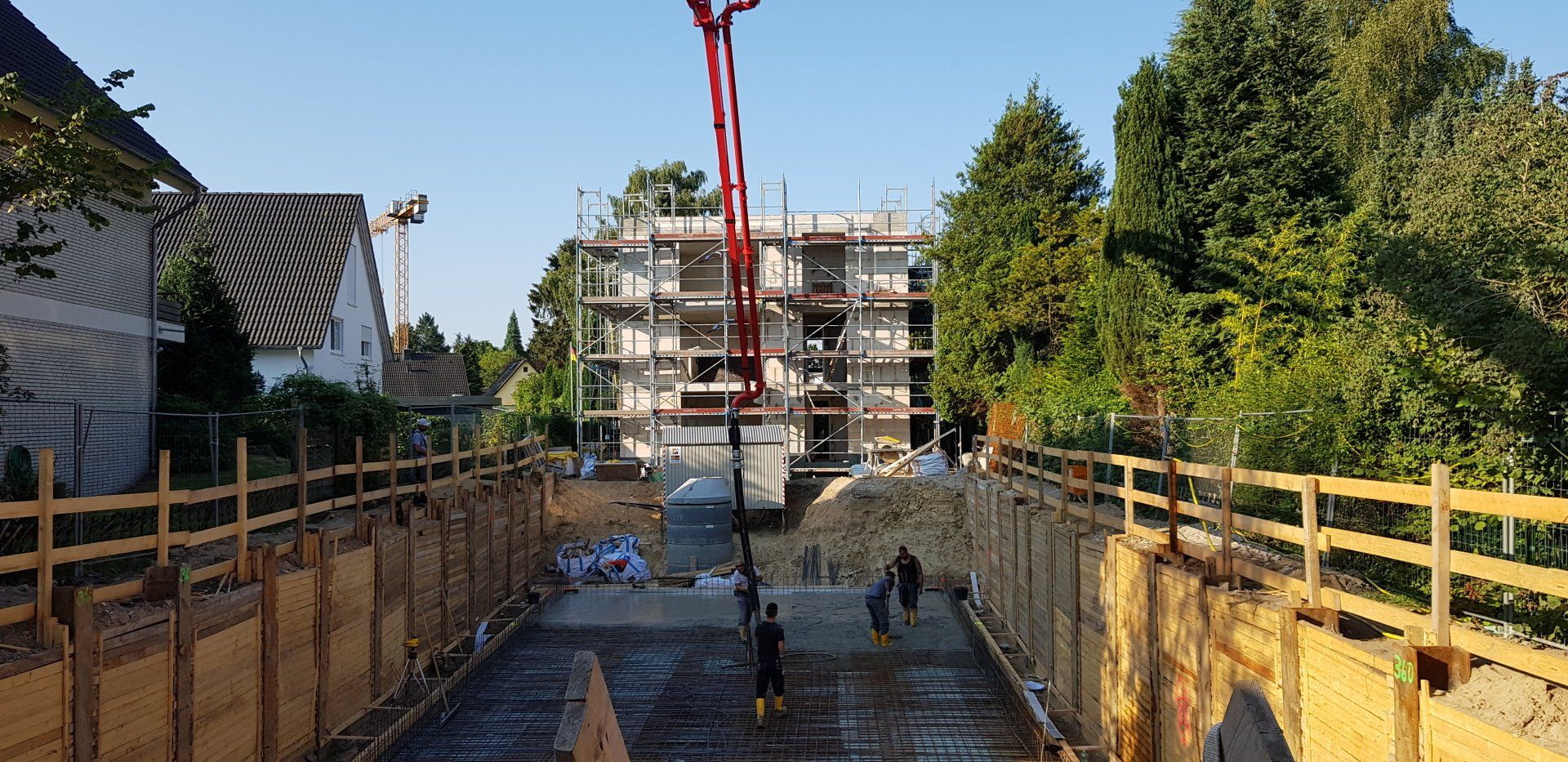 Baustelle mit Arbeitern auf einem Betonweg neben gelben Absperrungen und einem roten Kran, in der Nähe von Wohnhäusern