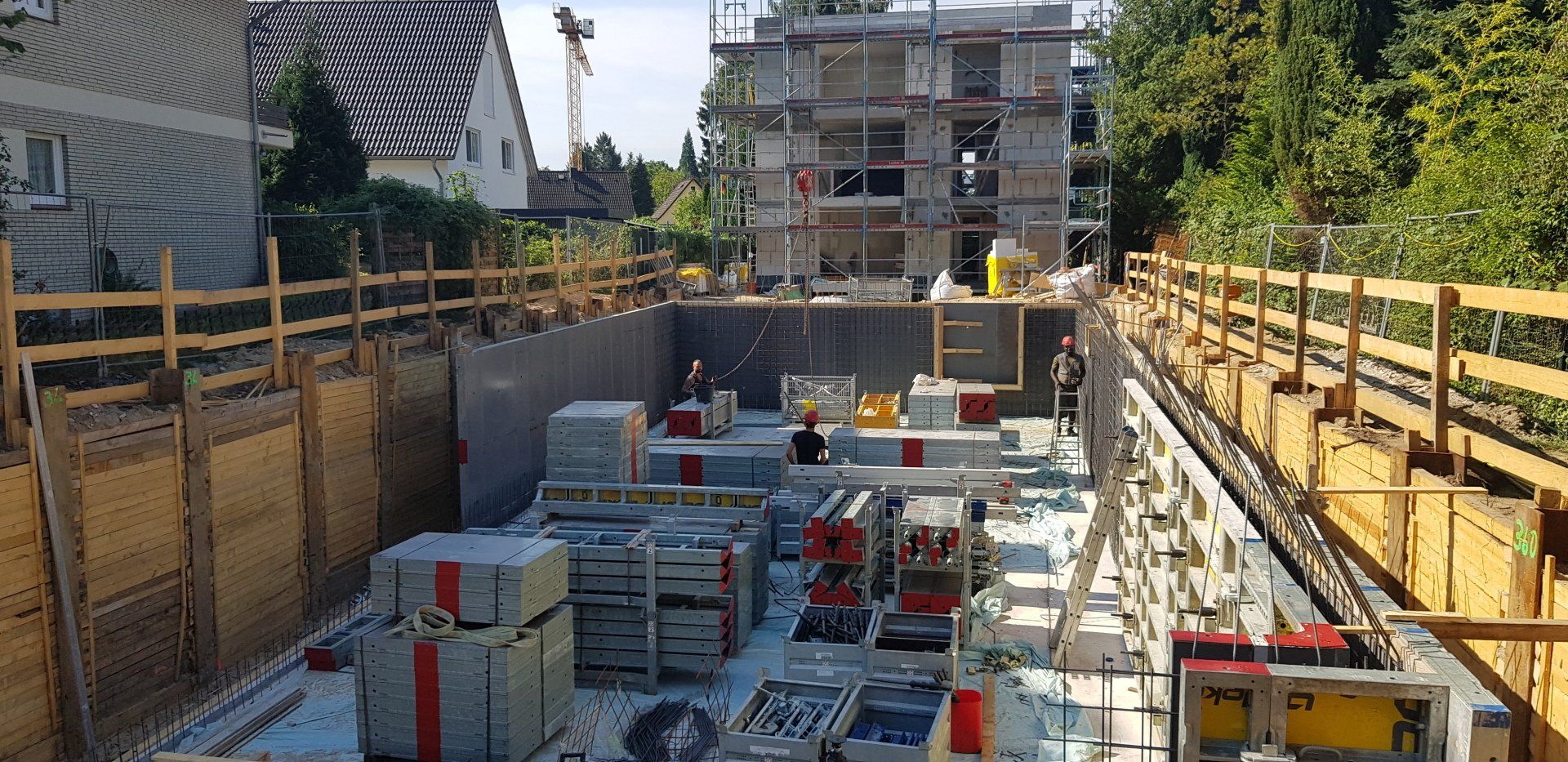 Baustelle mit einer tiefen Baugrube, gestapelten Materialien und Arbeitern unten neben einem eingerüsteten Gebäude