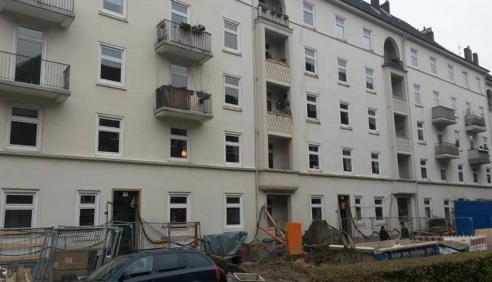 Weißes Mehrfamilienhaus mit Balkonen und Fenstern im Erdgeschoss, neben einer Baustelle mit Bauschutt und geparkten Autos