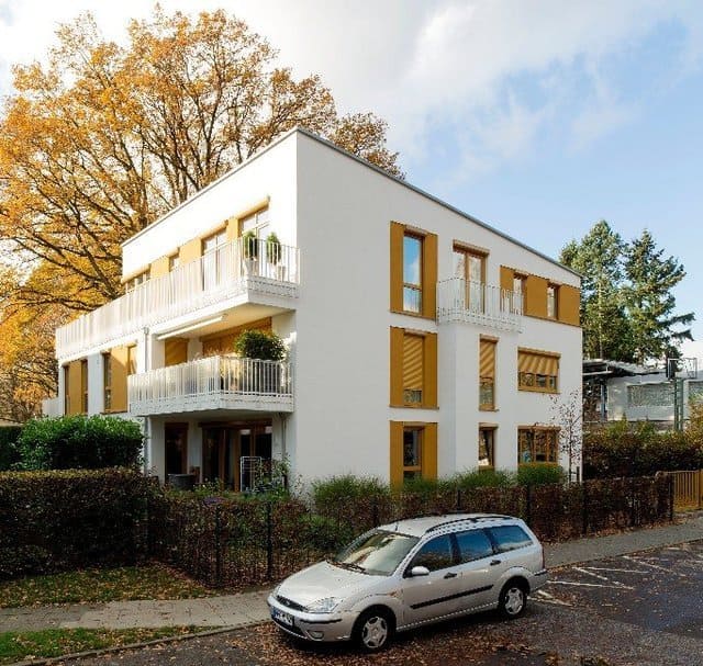 Modernes, weißes Mehrfamilienhaus mit Balkonen an einer baumbestandenen Straße, davor ein geparktes silbernes Auto.