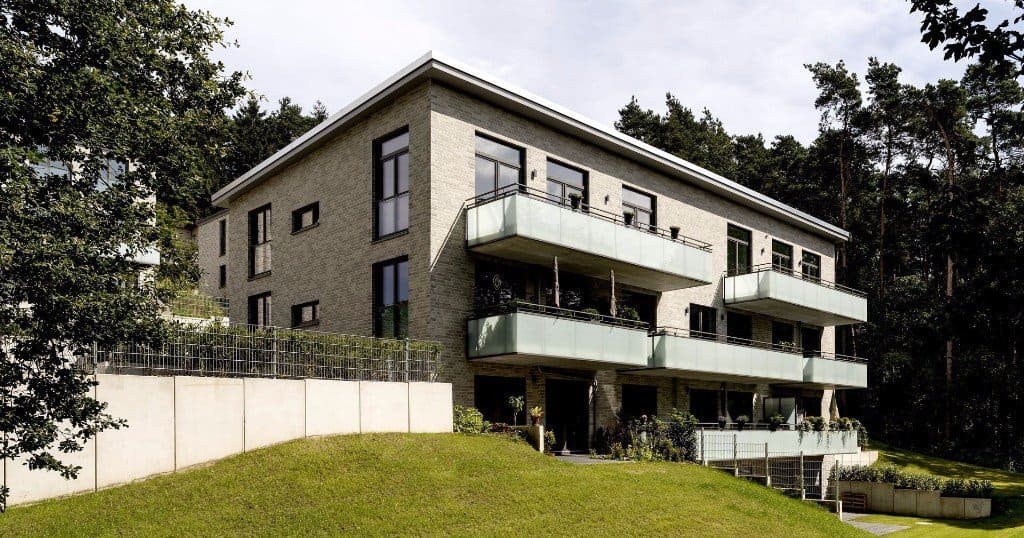 Modernes Mehrfamilienhaus mit Balkonen an einem grasbewachsenen Hang, umgeben von Bäumen und einem weißen Zaun