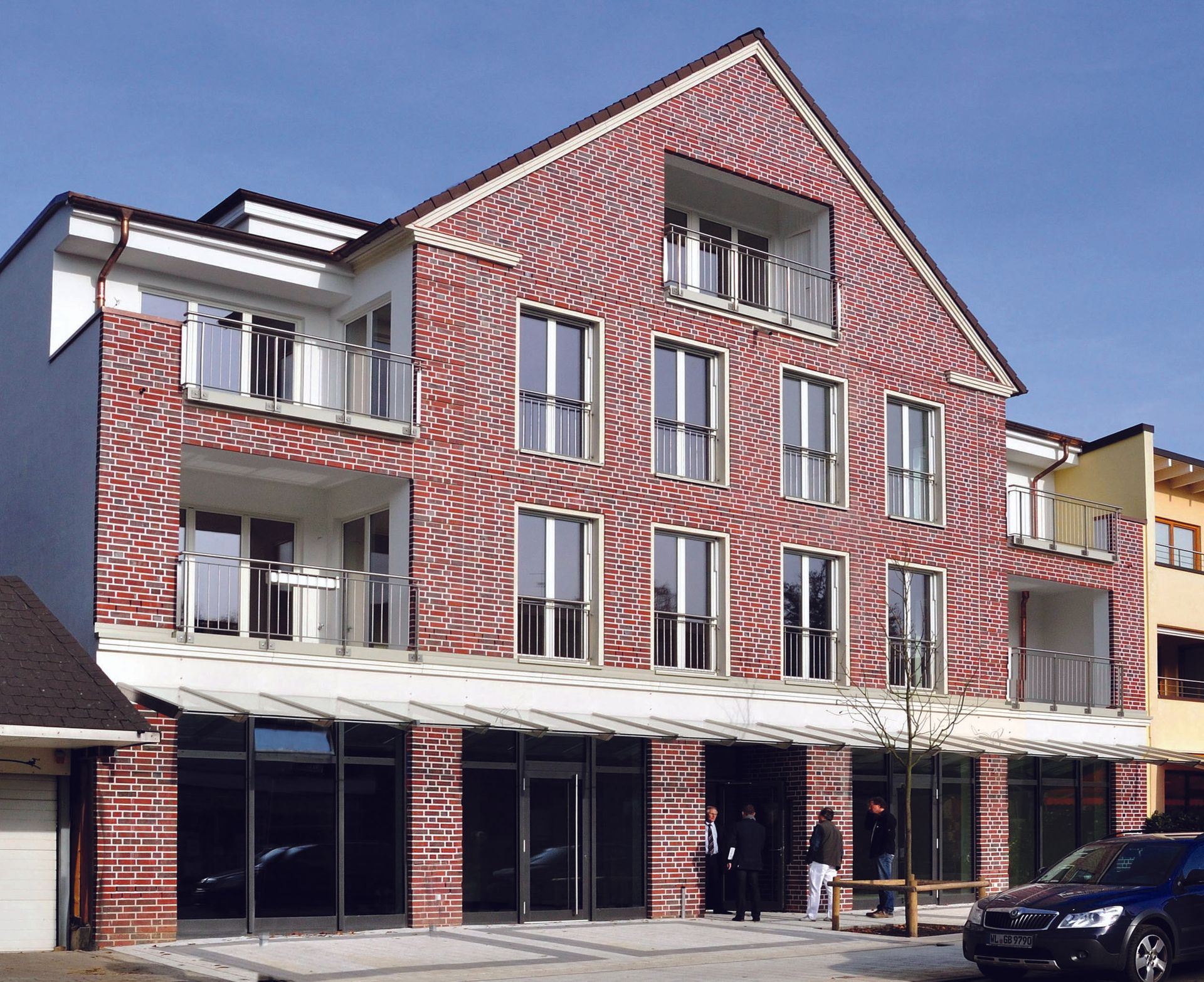 Ein mehrstöckiges Gebäude aus roten Backsteinen mit weißen Zierelementen und Ladenlokalen im Erdgeschoss an einer Straße
