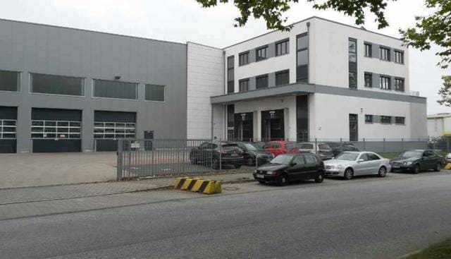 Modernes Industriegebäude mit davor geparkten Autos und einem Eingangstor unter bedecktem Himmel