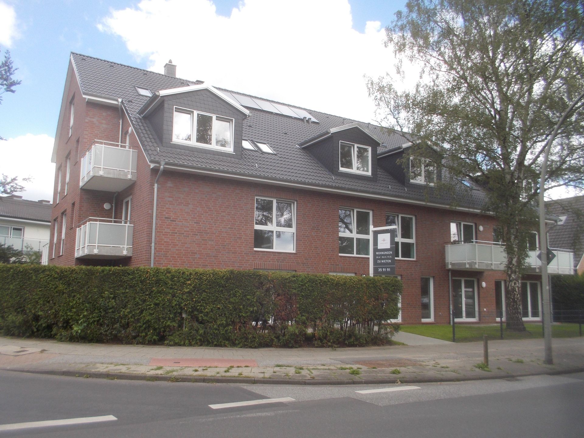 Dreistöckiges, rotes Backstein-Wohnhaus mit Balkonen, weiß gerahmten Fenstern und einer Hecke entlang der Straße