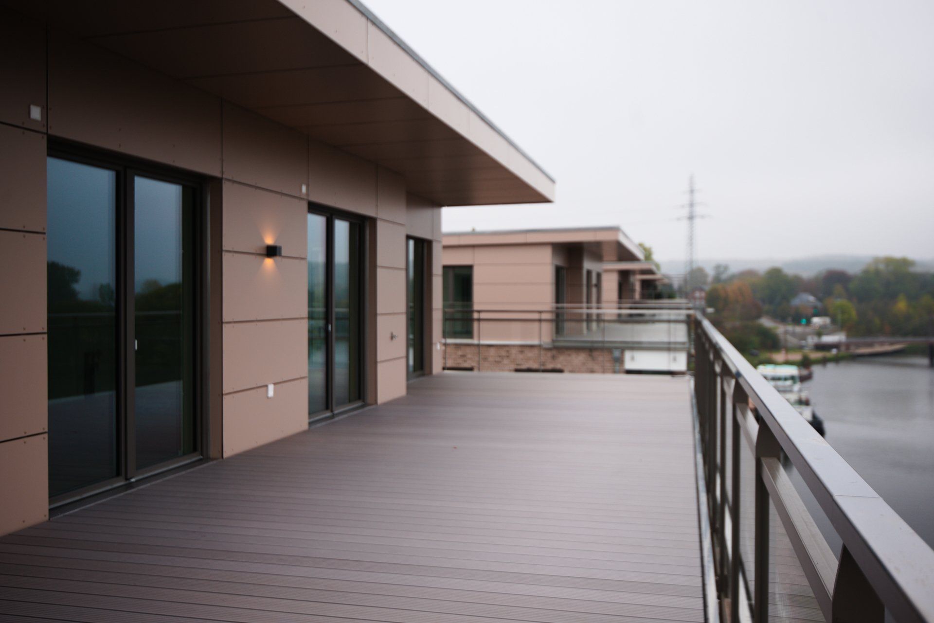 Moderne Dachterrasse mit Holzboden, Glasgeländer und beigefarbener Gebäudefassade mit Blick auf ein nebelverhangenes Wasser