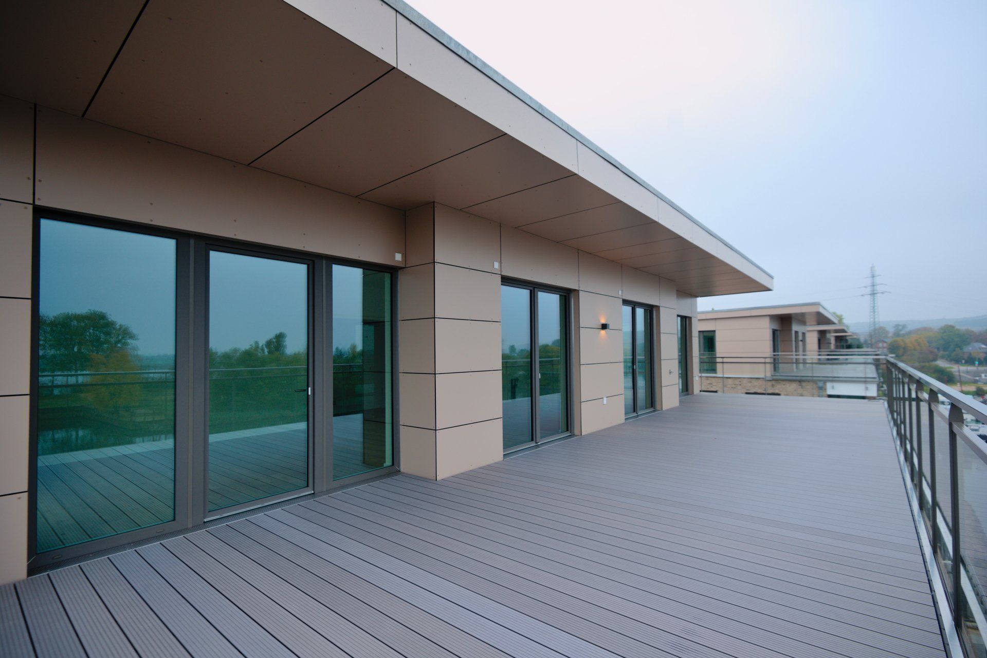 Moderne Gebäudeterrasse mit Glastüren, beigefarbenen Paneelen und einer breiten Holzterrasse mit Blick ins Grüne