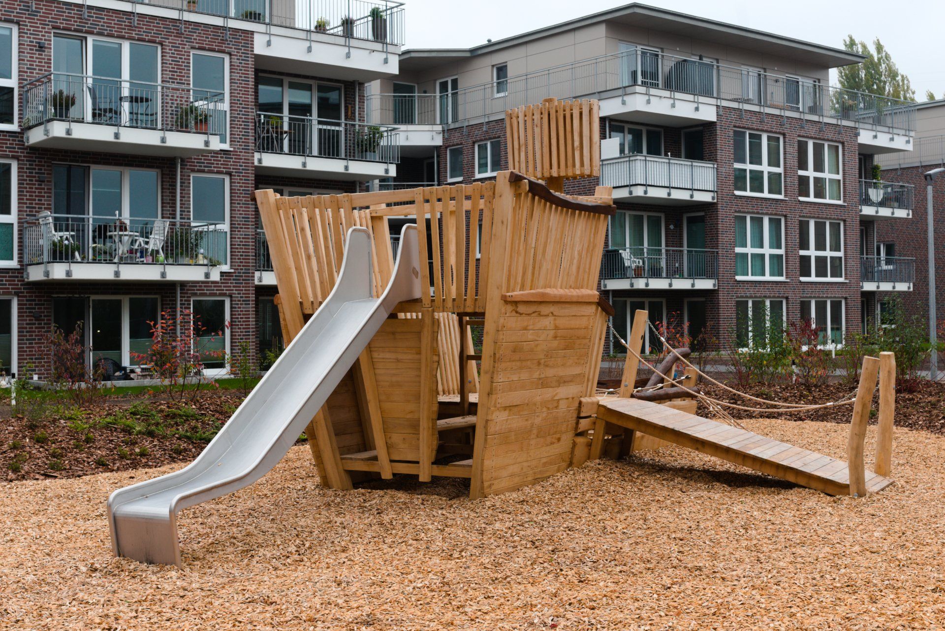 Spielplatzanlage aus Holz mit Rutschen in einem Innenhof, umgeben von Wohnhäusern.