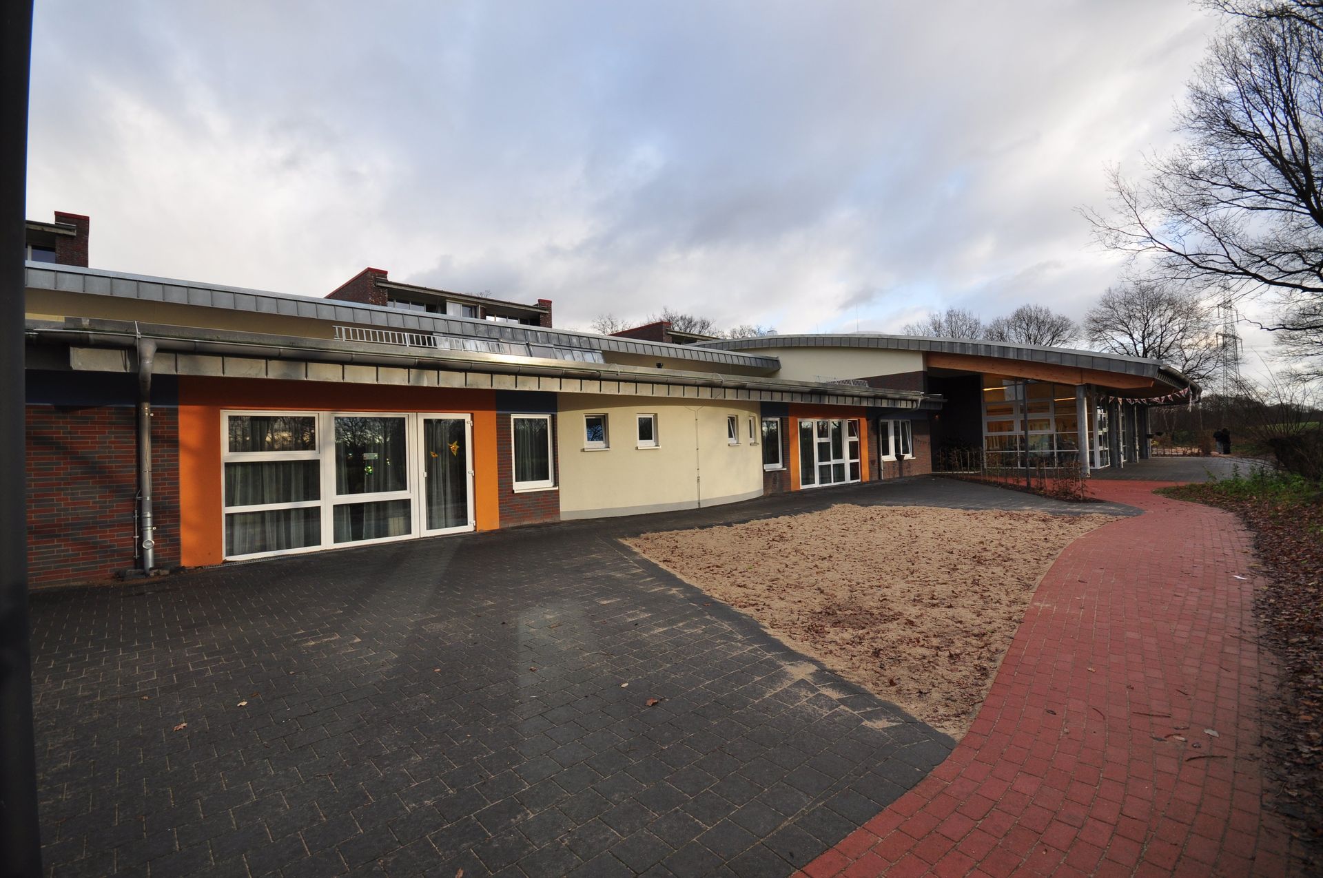 Eingeschossiges Gebäude mit großen Fenstern, schwarzer Auffahrt, Kieshof und rotem Weg unter bewölktem Himmel