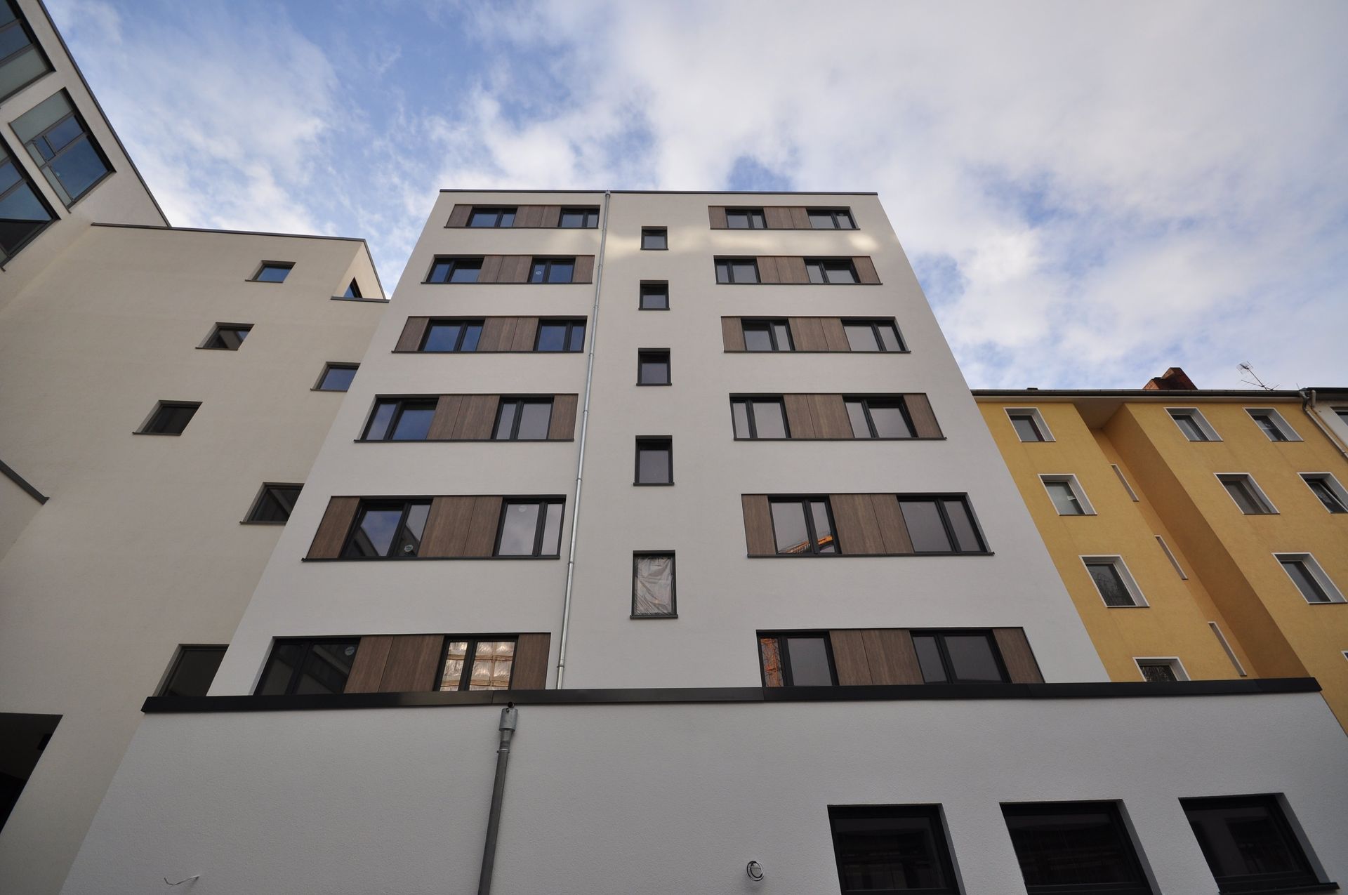 Ansicht von unten auf ein weißes Wohnhaus mit dunklen Fenstern neben gelben Gebäuden unter einem bewölkten Himmel