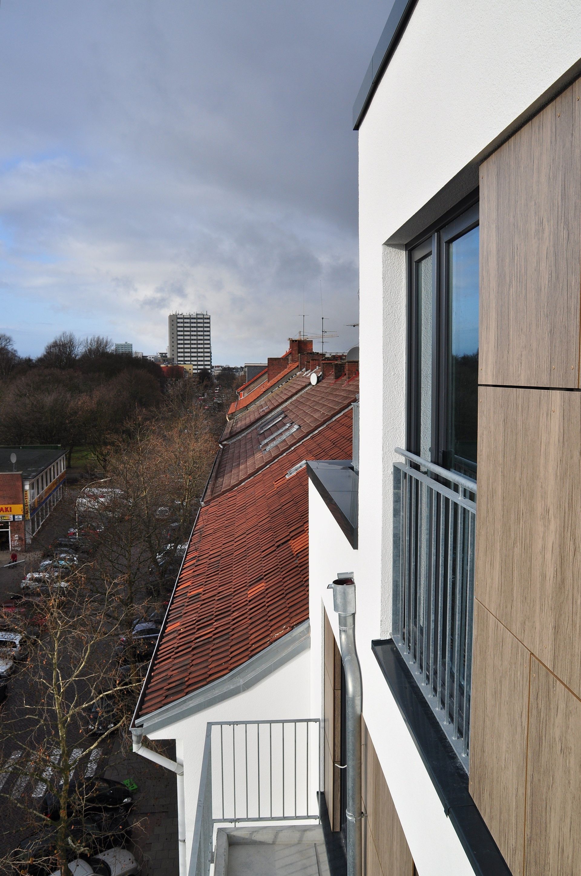 Blick vom Balkon über rote Dächer und eine Straße, im Hintergrund ein weißes Gebäude und bewölkter Himmel.