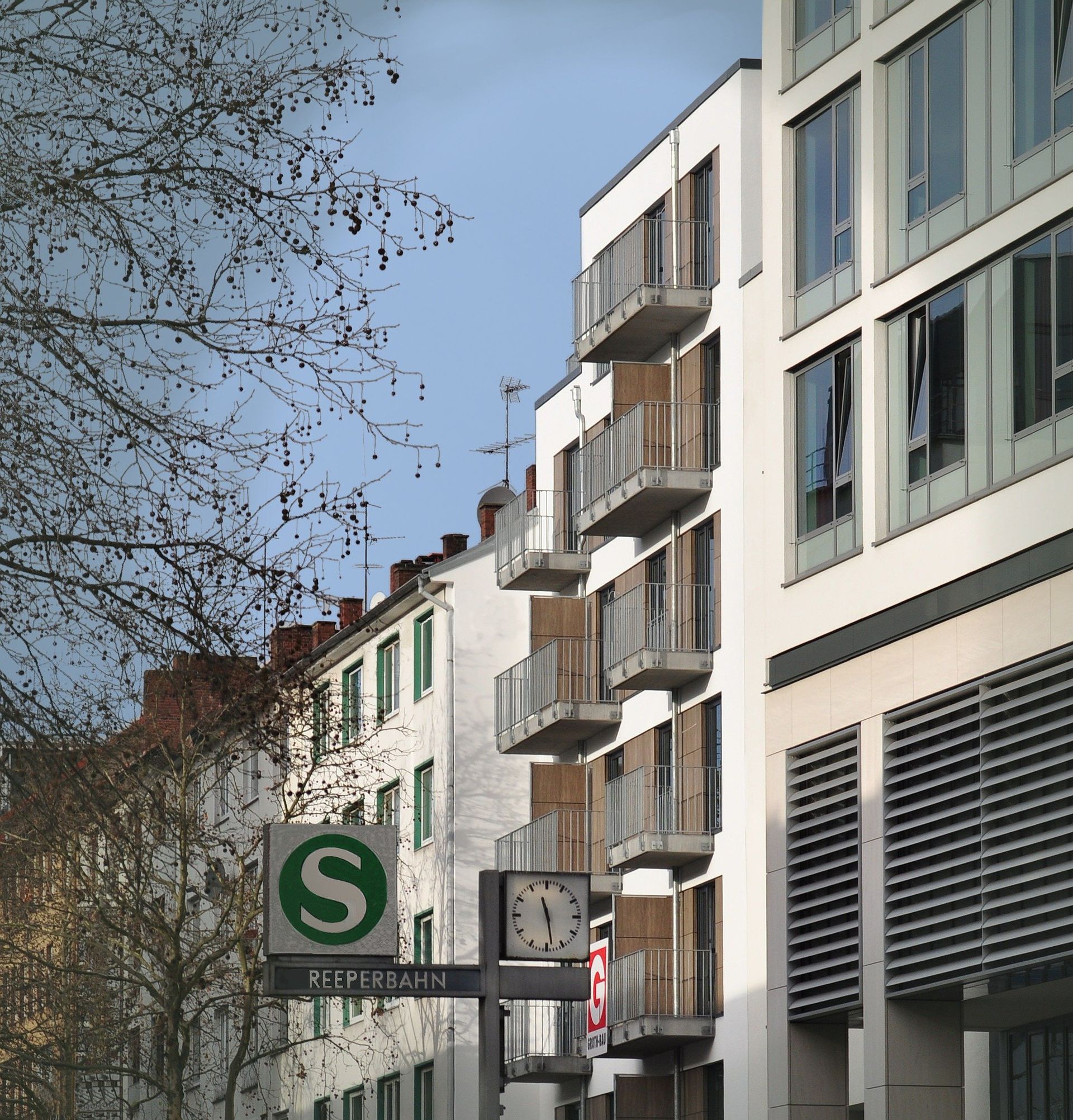 Eine städtische Straße mit einem grünen S-Schild, modernen Apartmentbalkonen und kahlen Bäumen unter blauem Himmel