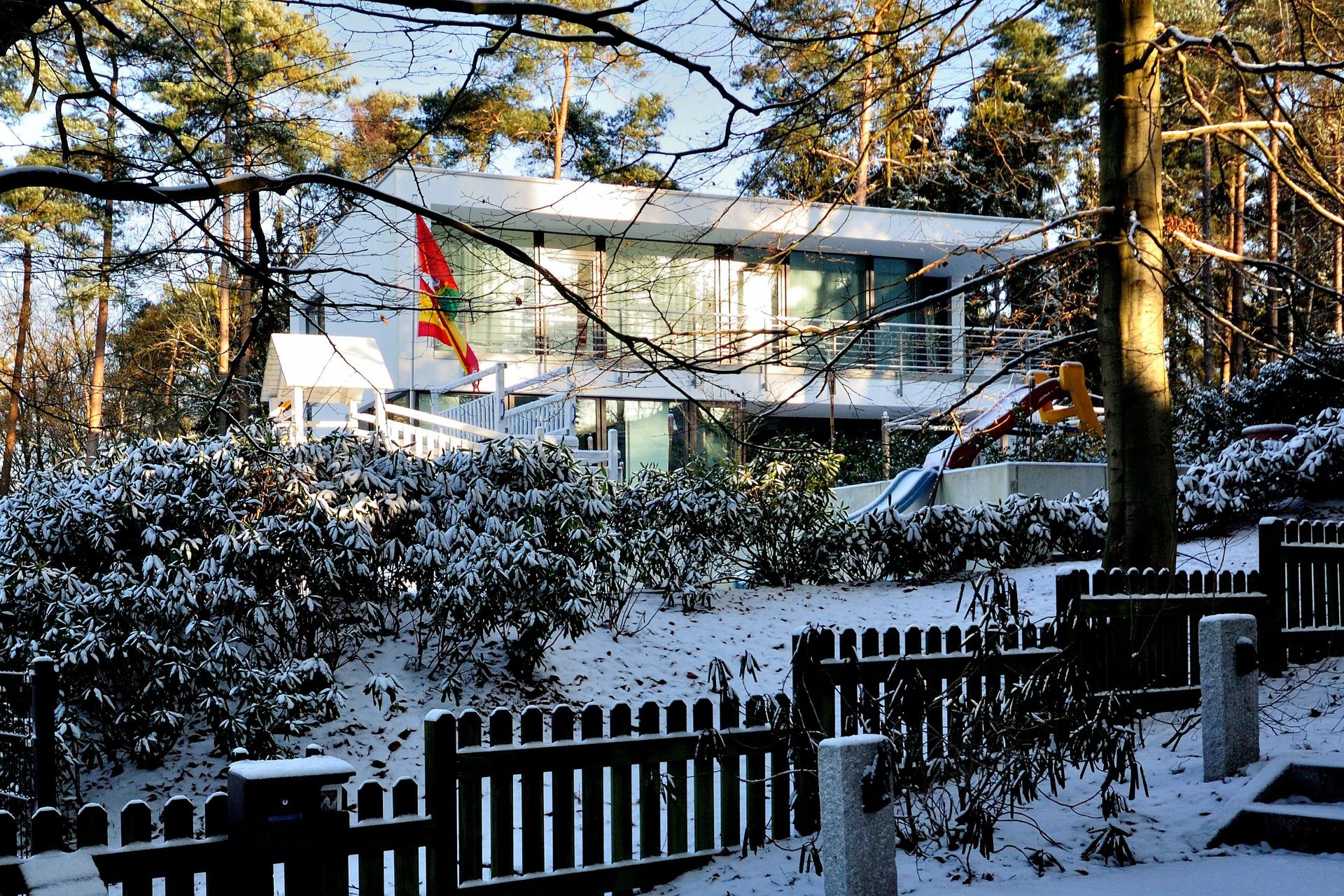 Ein schneebedecktes Haus hinter einem schwarzen Zaun, eingerahmt von hohen Bäumen in einer Winterlandschaft.