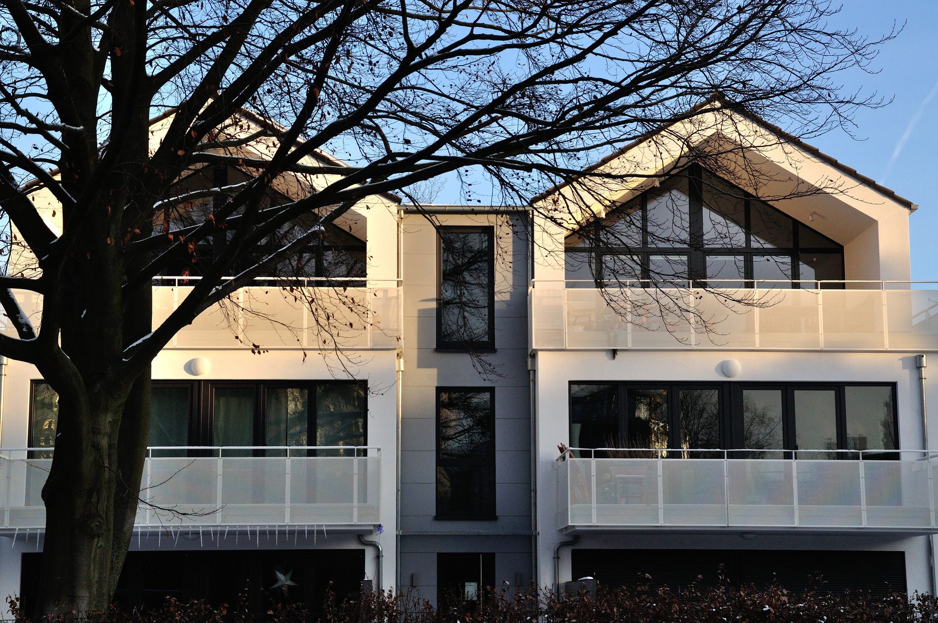 Zweistöckiges Haus mit Balkonen, einem zentralen Eingang und einem großen Baum davor